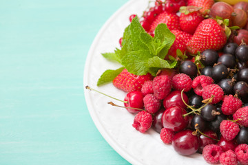 Closeup of white plate full of fresh organic berries: strawberries, blueberries, raspberries, gooseberry, blackcurrant decorated with mint as summer dessert or snack. Healthy food and eating concept