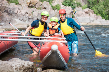 Young family of three wearing life vests and helmets posing beside rowing catamaran 