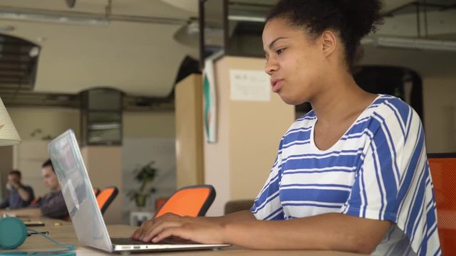 African young girl typing on the computer turns her head and smile. Near businesswoman sitting her colleagues at the work space. Mixed race employee wearing in casual top. Laughing sitting at