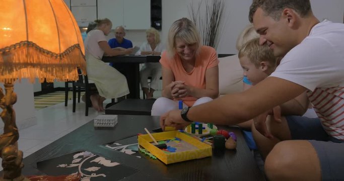 Child with father and two grandmothers playing with construction kits at home, other family members with pad in background