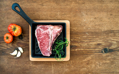 Raw uncooked meat t-bone steak with garlic, tomatoes, rosemary in small dark cooking pan over...