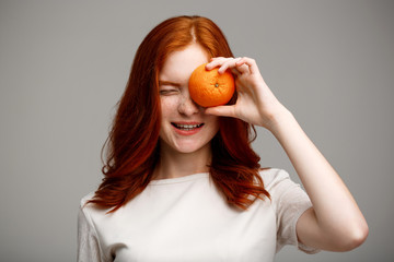 Portrait of beautiful ginger girl holding orange over gray background.