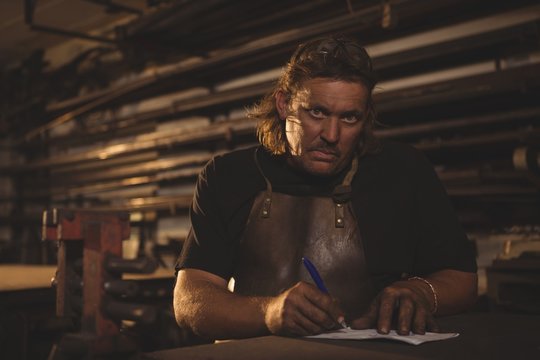 Portrait of blacksmith preparing notes while sitting in workshop - Powered by Adobe