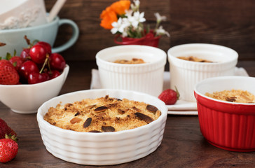 Cherry, strawberry crumble dessert in white bowls. Cherries, strawberries, flowers behind. Dark wood background, top view