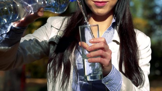 Slowmotion young beautiful happy woman stands in park and pours water from plastic bottle to glass