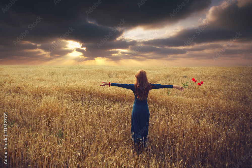 Wall mural Back view of woman who stands toward the sunset in a wheat field with arms spread out, holding a poppy flower - Wall murals