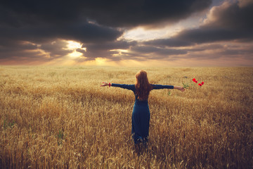 Back view of woman who stands toward the sunset in a wheat field with arms spread out, holding a poppy flower