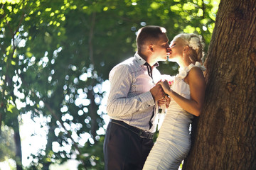 Fototapeta premium Kiss in the sunlight - groom kisses a bride leaning to a tree