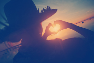 Naklejka premium Woman in straw hat making heart symbol with her hands at sunset (vintage color)