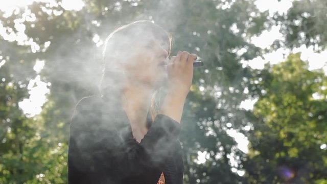 Girl smokes electronic cigarette sitting on the parapet in the park close-up