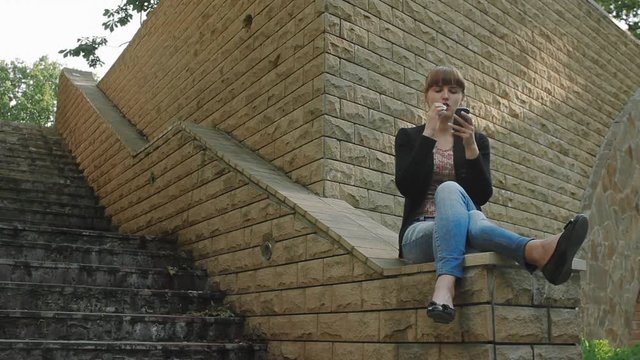 Girl smokes electronic cigarette sitting on the parapet in the park and surfing social net