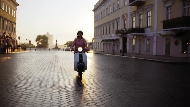 Young beautiful girl riding around city on scooter at sunrise. Slow motion.