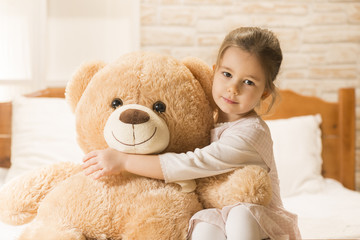 Sweet little girl is hugging a teddy bear, looking at camera and smiling while sitting on her bed at home