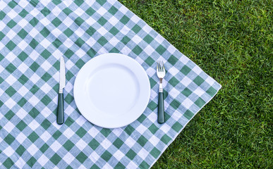 Knife, white plate and fork on green picnic tablecloth. Flat lay
