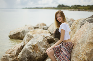 young beautiful girl sitting on the rocks by the sea with a black hat in hands