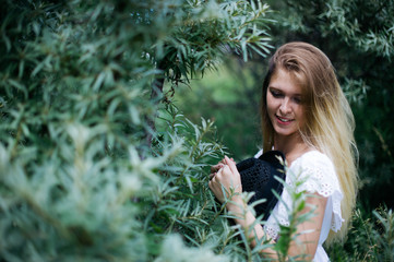 young beautiful girl in a white dress with a black hat looks out from behind a bush