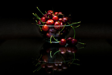 Transparent cup with red ripe cherries with reflection on black background.