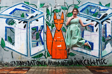 Naklejka na ściany i meble Young happy girl in a dress jumping, background graffiti