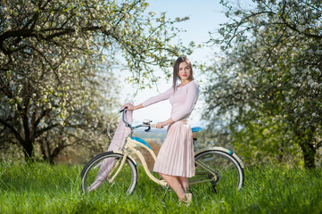 Beautiful young woman with long hair wearing nice white dress keeps retro bike against the background of flowering trees and fresh greenery in spring garden