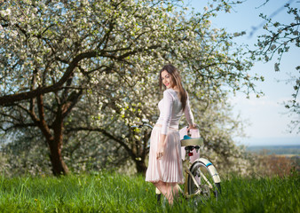 Fototapeta premium Beautiful brunette woman wearing a nice white dress with bike standing with her back and turned to the camera in a fresh greenery of spring garden