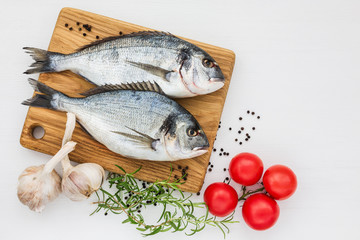 Fresh dorado fish on wooden cutting board with garlic and tomatoes on white table. Top view, copy space.