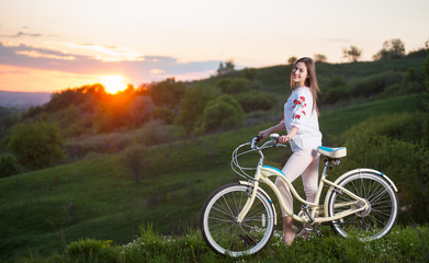 Obraz premium Happy girl with retro bike standing at hill and looks to the camera, with a blurred background of greenery and beautiful sunset