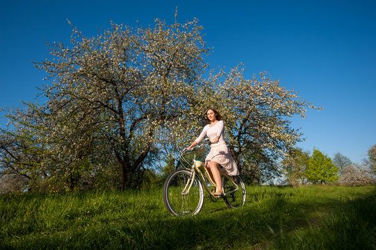 Smiling brunette woman wearing light dress riding a vintage white bicycle against blooming trees, fresh greenery and blue sky in the spring garden at the sunny day. Wide angle lens