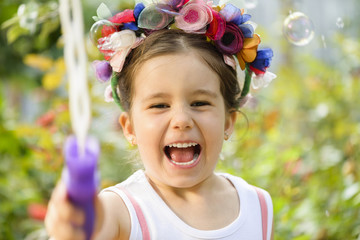 Happy little girl wearing flower wreath playing outdoor with soap balloons in a sunny summer day
