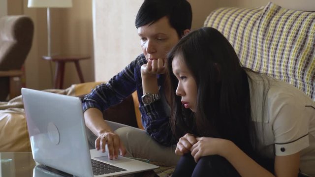 Adult caucasian woman looking at laptop explain something young asian girl. Multicultural team work with computer indoors. Vietnamese girl focus on screen. Older female shows errors that must be