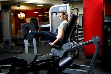 An attractive girl working our on a huge training simulator in a gym.Sportive woman using weights press machine for legs at gym.Pretty brunette exercising in simulator. Working her quads at machine.