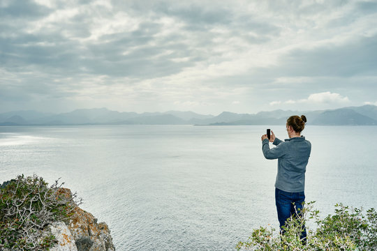 Young man standing on cliff photographing with smartphone, Alcudia, Majorca, Spain