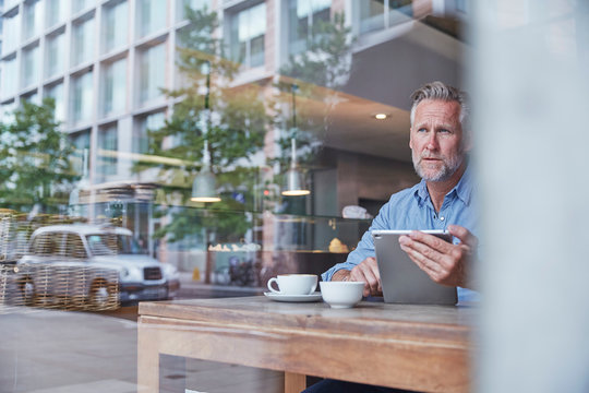 Mature man sitting in cafe, using digital tablet, street reflected in window