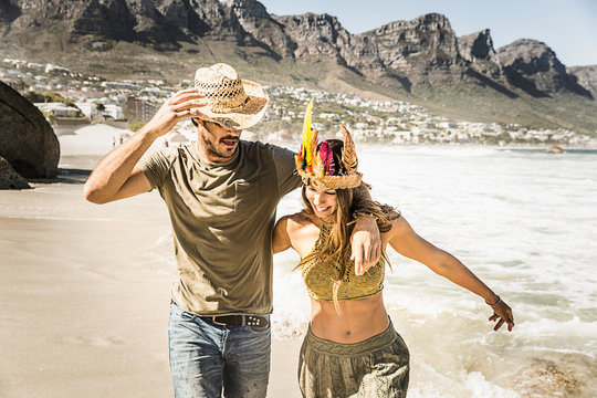 Mid adult couple wearing straw hat and feather headdress running on beach, Cape Town, South Africa