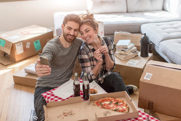 Moving house: Young couple taking self portrait with smartphone, in new home, surrounded by boxes