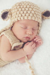 Cute newborn baby knitted in beige overalls and a beige knitted cap with round ears, folded handle under the head and legs legs, happily sleeping on a white, fluffy, soft blanket 