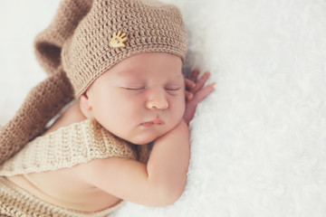 Cute newborn baby in a beige knit suit and a brown knitted cap, folded under the handle head and pursing his feet, happy to sleep on a white, fluffy, soft blanket