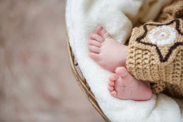 Parts of a body - pink legs of the newborn child sleeping on a white blanket in a round basket on a brown background, covered with a knitted brown blanket