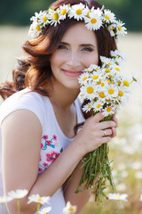 Cute pregnant brunette woman in white t-shirt with a bouquet of white daisies on his head wears a wreath of white flowers,chamomile,summer,walking alone in a beautiful field of blooming white daisies
