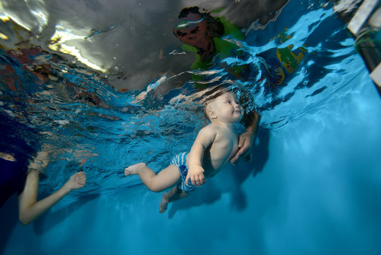 Happy baby trains and swims underwater in the pool on a blue background, and the dad catches him. Bottom view. Horizontal orientation