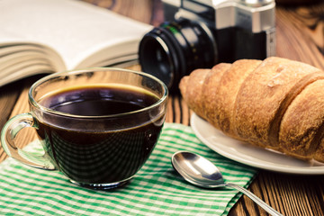 Cup of black coffee on napkin with croissant, open book, vintage camera, wooden table, travel concept