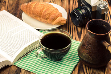 Cup of black coffee on napkin with croissant, open book, turkish coffee pot, vintage camera, wooden table