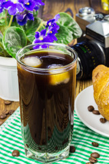 Glassful of black coffee with ice on green napkin. croissant, vintage camera, flowerpot, wooden table, selective focus