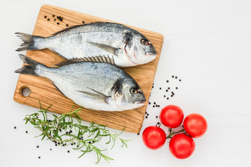 Fresh dorado fish on wooden cutting board with tomatoes on white table. Top view, copy space. 