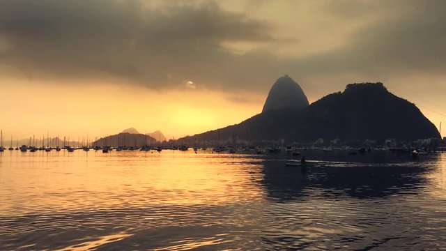 A lone rower passes in front of a golden sunrise silhouette of Sugarloaf Mountain at Botafogo Bay in Rio de Janeiro, Brazil