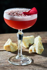 Alcoholic cocktail raspberry sauer consisting of liqueurs, vermouth, chocolate chip and lemon juice served in a glass on wooden table on black background