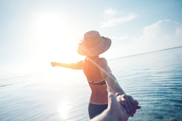 Handsome woman in hat guiding a man by the hand into the ocean (intentional sun glare) - obrazy, fototapety, plakaty