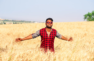 Young indian man walking in wheat field - African american guy open arms at sky through a corn field - Asian farmer with open arms towards the sun - Concept of freedom and joy for successful achieved
