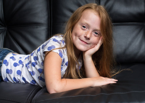 Little ginger girl with freckles is laying on the black sofa, head is leaning on the hand