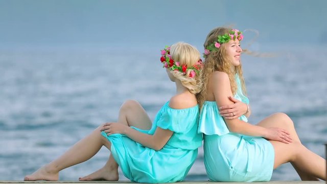 Cute girls with wreaths on their heads are resting on the river. Two young beautiful woman in a blue dress sits on a wooden pier on the river.