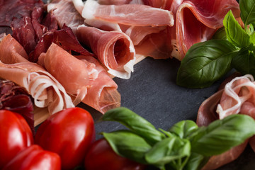 Jambon mix. Ham. Traditional Italian and Spanish salting, smoking, dry-cured dish - jamon Serrano and prosciutto crudo sliced with herbs and tomatos on dark stone background. Copy space. Closeup. 
